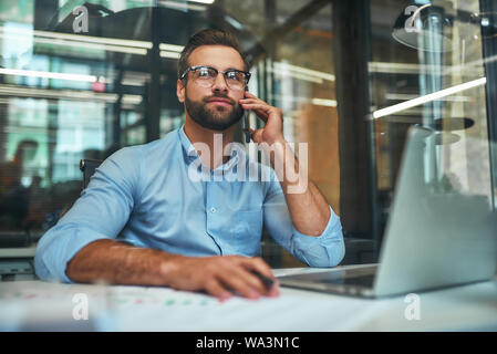 Heureux de vous aider. Portrait de jeune homme séduisant dans les lunettes et l'usure formelle à parler au téléphone avec le client tout en restant assis dans le bureau. Lieu de travail. Concept d'affaires Banque D'Images