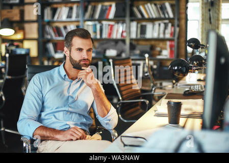 Bonne journée de travail. Jeune homme barbu dans l'usure formelle de toucher son menton et penser à quelque chose alors qu'il était assis dans un bureau moderne. Lieu de travail. Concept d'affaires Banque D'Images