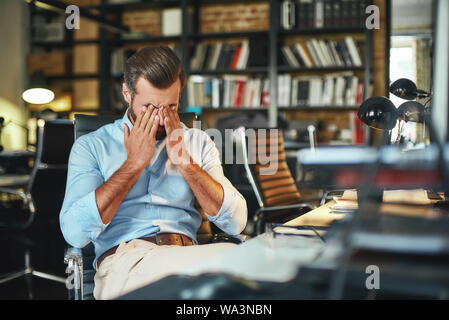 Travail dur. Jeune homme barbu épuisé couvrant ses yeux et stressé alors qu'il était assis dans un bureau moderne. Lieu de travail. Concept d'affaires Banque D'Images