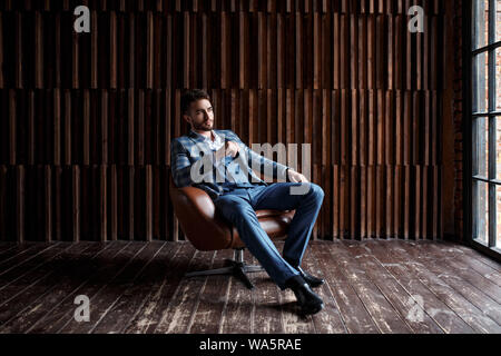 Jeune mec avec une barbe dans un costume bleu est assis sur une chaise et regarde dans la distance Banque D'Images