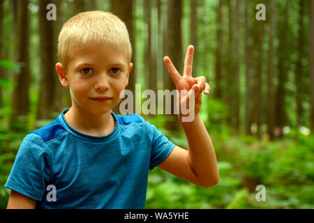 L'été lumineux portrait of a 6 ans vilain garçon caucasien blond faisant signe V de la victoire avec ses doigts et smiling over woody background Banque D'Images