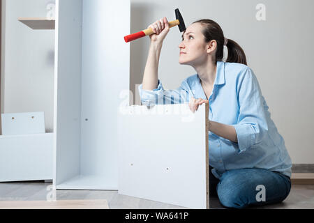 Portrait d'une jeune femme blanche avec un marteau à la main assis sur le plancher et collecte des meubles en bois blanc Banque D'Images