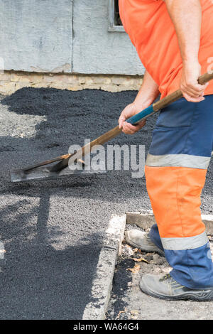 L'homme travailleur fixe la route d'asphalte pavage réparation soleil jaune lumineux. Un homme en salopette est de jeter l'asphalte avec une pelle. Photo verticale. Banque D'Images