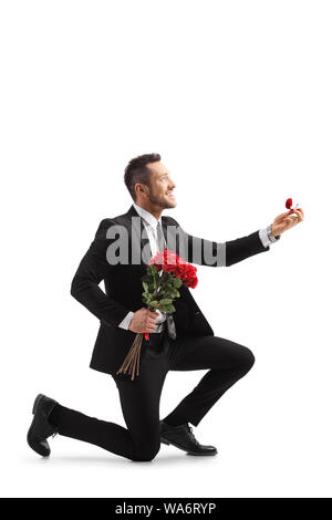 Jeune homme élégant dans un costume à genoux et tenant des roses et une bague de fiançailles isolé sur fond blanc Banque D'Images