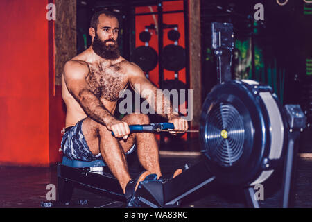 Jeune homme barbu à l'aide d'une machine à ramer dans la salle de sport Banque D'Images