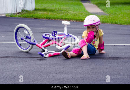 Petite fille est assise sur le sol en Casque et équipement de sécurité, après avoir chuté de son vélo, tout en apprenant à faire avec les roues de formation Banque D'Images