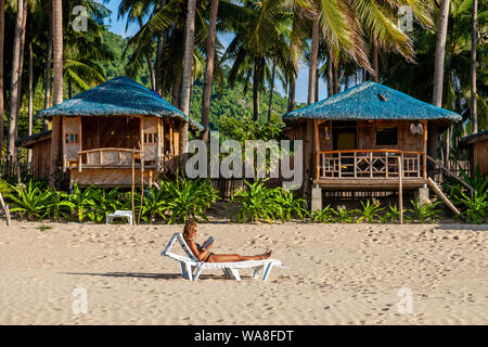 Une femme touriste se prélasse sur un transat en lisant un livre à l'extérieur de son hébergement en bord de mer, Nacpan Beach, El Nido, l'île de Palawan, aux Philippines. Banque D'Images