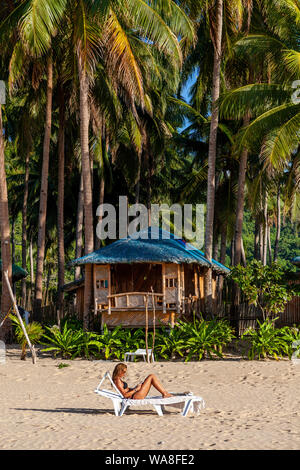 Une femme touriste se prélasse sur un transat en lisant un livre à l'extérieur de son hébergement en bord de mer, Nacpan Beach, El Nido, l'île de Palawan, aux Philippines. Banque D'Images