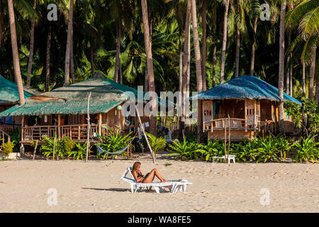 Une femme touriste se prélasse sur un transat en lisant un livre à l'extérieur de son hébergement en bord de mer, Nacpan Beach, El Nido, l'île de Palawan, aux Philippines. Banque D'Images