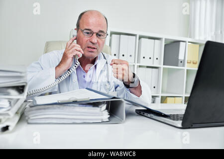Mature en colère médecin chef à la recherche de document dans le dossier ouvert sur sa table et chouting au récepteur du téléphone Banque D'Images