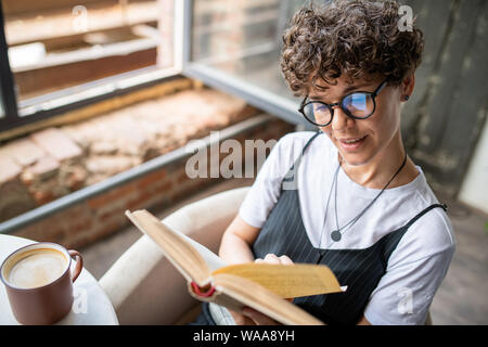Jeune femme avec de courts cheveux bruns bouclés et la lecture de livre à la maison Banque D'Images