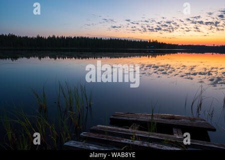 De soleil colorés et de la solitude dans un lac finlandais avec de belles réflexions des nuages et sombre forêt boréale et ancienne jetée en premier plan, Finlan Banque D'Images