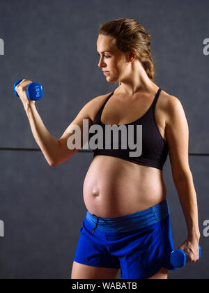 Femme enceinte d'une trentaine d'exercer dans une salle de sport faire des biceps avec haltères. Banque D'Images