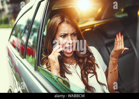 Les jeunes en colère business woman holding smartphone est dans sa main et fait valoir avec quelqu'un, sur le siège arrière de la voiture. Banque D'Images