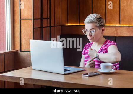 Date limite ! Portrait of attractive young avertissement bossy girl cheveux courts est assis dans un café et faire appel vidéo sur ordinateur portable et montrant checki Banque D'Images