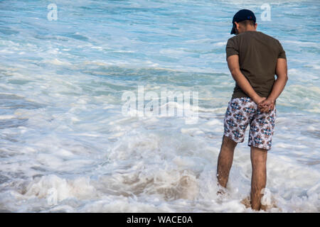 Un homme en vêtements décontractés pensivement standing in ocean avec les mains derrière le dos tout en regardant vers le bas dans une profonde réflexion. Les vagues éclaboussant sur lui Banque D'Images