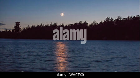 Un croissant de lune illuminée par le soleil couchant jette une lumière dorée sur un lac du nord du Wisconsin. Banque D'Images