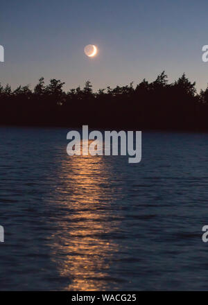 Un croissant de lune illuminée par le soleil couchant jette une lumière dorée sur un lac du nord du Wisconsin. Banque D'Images