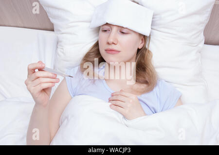 Froid et mal de tête. Caucasian girl couchée sur le lit avec un thermomètre dans la main et une serviette humide sur son front. Banque D'Images