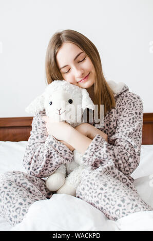 Jeune femme aux cheveux blonds assis sur le lit dans sa chambre à coucher dans une étreinte avec un animal en peluche jouet. Moutons préférés dans les mains de teen girl Banque D'Images