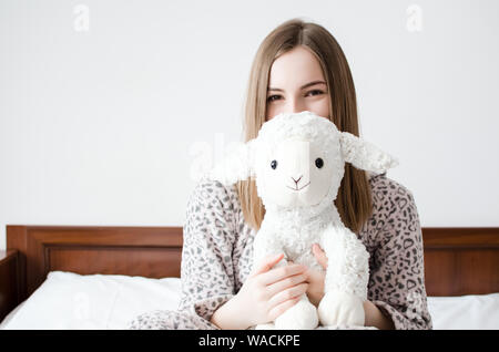 Jeune femme aux cheveux blonds assis sur le lit dans sa chambre à coucher dans une étreinte avec un animal en peluche jouet. Moutons blancs préférés dans les mains du t Banque D'Images