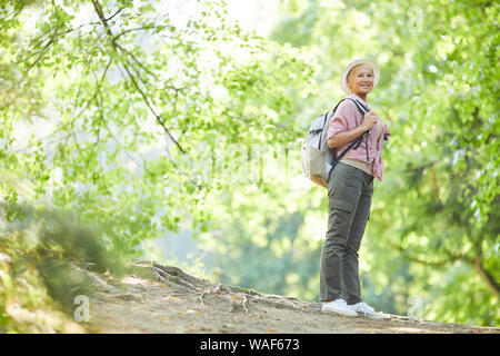 Young woman in casual clothing avec sac à dos derrière son dos standing and smiling au cours de sa promenade dans la forêt en été Banque D'Images