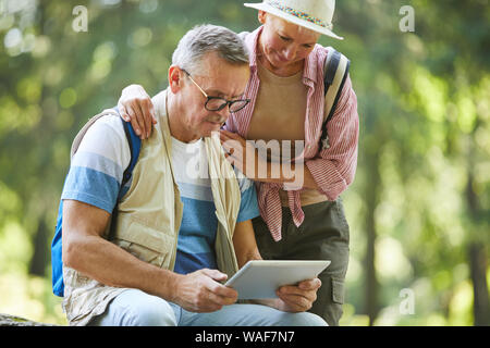 Senior man wearing eyeglasses sitting et regarder quelque chose sur tablette numérique avec des hauts femme debout près de lui pendant qu'ils sont dans la forêt Banque D'Images