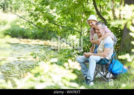 Mature Woman embracing l'homme alors qu'il assis sur une chaise et la pêche au bord de lac Banque D'Images