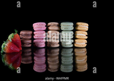 Délicieux dessert macaron en différentes couleurs - empilés à côté de macarons deux fraises rouges juteux sur une table en verre et un fond noir - Close-up Banque D'Images