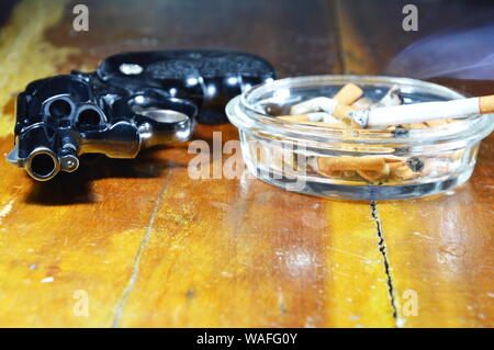 Pistolet revolver et cigarette dans le cendrier en verre table en bois Banque D'Images