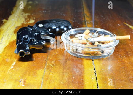 Pistolet revolver et cigarette dans le cendrier en verre table en bois Banque D'Images