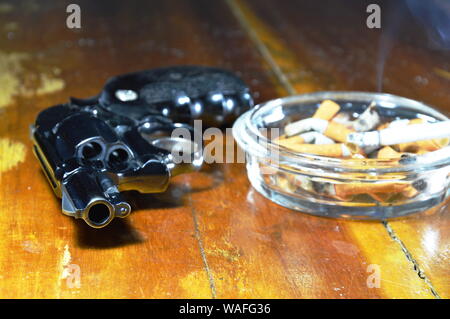 Pistolet revolver et cigarette dans le cendrier en verre table en bois Banque D'Images