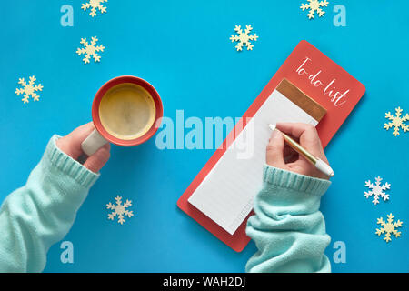 N'Noël liste et woman's hands en couleur menthe pullover avec tasse de café sur la lumière bleu pastel table. Mise à plat, de style vue d'en haut. La planification de vacances. Banque D'Images