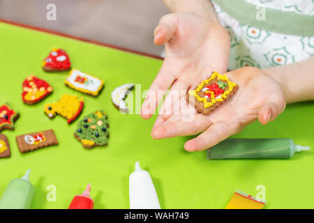 Les mains des enfants sont maintenant un gâteau rectangulaire décoré. Cannelle gâteaux colorés se trouvent sur la table verte à l'arrière-plan. Tubes avec glaçage coloré Banque D'Images