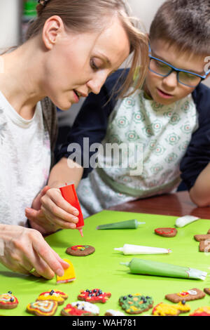 Mère et son fils dans la cuisine. Une femme décore un gâteau à la cannelle avec un tube de rouge cerise. Le fils observe attentivement ce que sa mère est en train de faire. De Banque D'Images