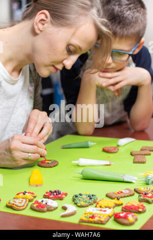 Mère et son fils dans la cuisine. Une femme décore un gâteau à la cannelle avec un tube de rouge cerise. Le fils observe attentivement ce que sa mère est en train de faire. De Banque D'Images