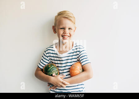 Portrait garçon blond dans le contexte d'un mur blanc détient deux citrouilles sous les aisselles et rit. Banque D'Images