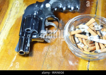 Pistolet revolver et cigarette dans le cendrier en verre table en bois Banque D'Images