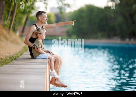 Photo de père et boy sitting on wooden bank par jour d'été sur la rivière Banque D'Images