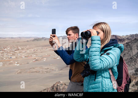 La Mongolie Ulgii 2019-05-04 fille européenne regardant à travers des jumelles et jeune homme faire de photos sur l'appareil photo du smartphone. Sur la montagne à Concept backpackers Banque D'Images