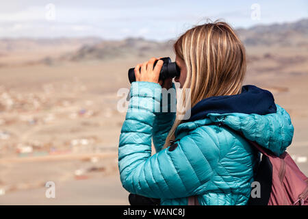 La Mongolie Ulgii 2019-05-04 European blonde girl in blue Jacket regardant à travers des jumelles, randonnées, escalade, marche sur fond de montagne. Concept lon Banque D'Images