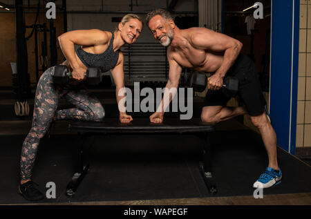 A middle aged couple attrayant et mettre en place est en train de faire un entraînement de fitness fonctionnel avec haltères. Homme fort et la femme sont en train de rire dans une salle de sport. Banque D'Images