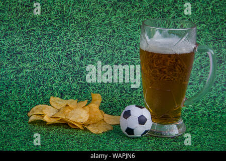 Verre à bière avec de la bière, chips, ballon de football sur un fond d'herbe verte Banque D'Images