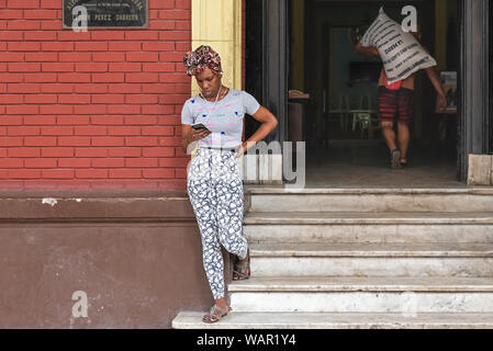 La Havane, Cuba - 09 avril, 2019 : une femme regarde son téléphone portable tout en se tenant à l'extérieur et l'entrée de l'édifice dans la vieille ville Banque D'Images