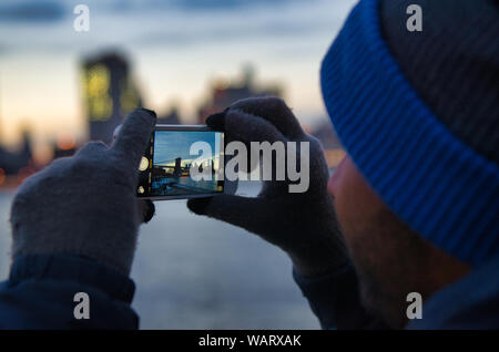En hiver homme chapeau et mitaines prend une photo du coucher de soleil derrière le pont de Brooklyn, par une froide soirée d'hiver. Banque D'Images