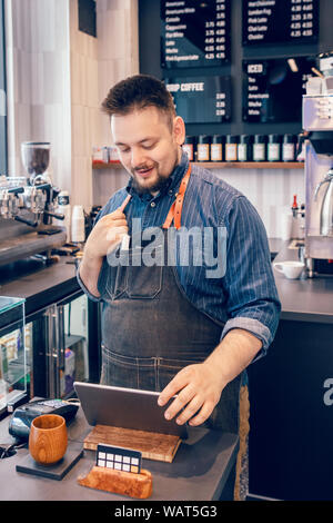 Jeune homme caucasien homme barbu en restaurant café barista caissier au travail. Vendeur sur le bloc de touches pour l'acceptation de paiement client client achat. Sm Banque D'Images