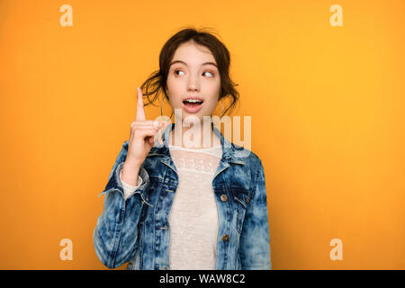 Une jeune fille brune en jeans se dresse sur un fond jaune et avec enthousiasme sur le côté, soulève l'index de la page Banque D'Images