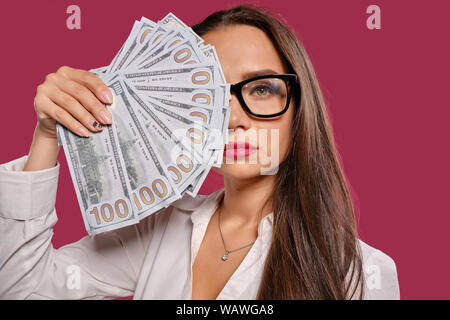 Fille brune à lunettes, vêtu d'une jupe courte noire et chemisier blanc est posing holding a fan of hundred dollar bills contre un fond rose. Banque D'Images
