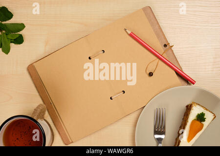 Un ordinateur portable ouvert de papier kraft avec un crayon rouge se trouve sur une table en bois clair avec une plaque avec un dessert et une tasse de thé. Banque D'Images