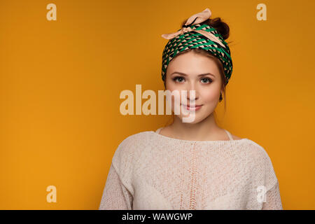 Une jeune fille dans un pull léger et un foulard noué est souriant debout sur un fond jaune. Banque D'Images